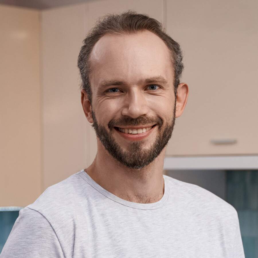 Homme souriant avec une barbe, portant un t-shirt blanc dans un intérieur lumineux